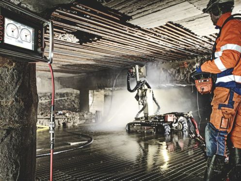 Trabalhador opera máquina de hidrodemolição em túnel com teto de concreto, usando uniforme laranja e equipamento de proteção.