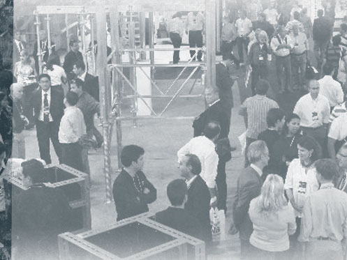 Pessoas circulam e conversam em evento ou feira industrial, cercadas por estruturas metálicas e estandes. Imagem em tons de cinza.